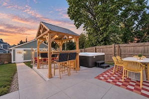 Ein Garten mit einer Holzw pergola, Sitzgelegenheiten im Freien, einem Whirlpool und einer karierten Matte.