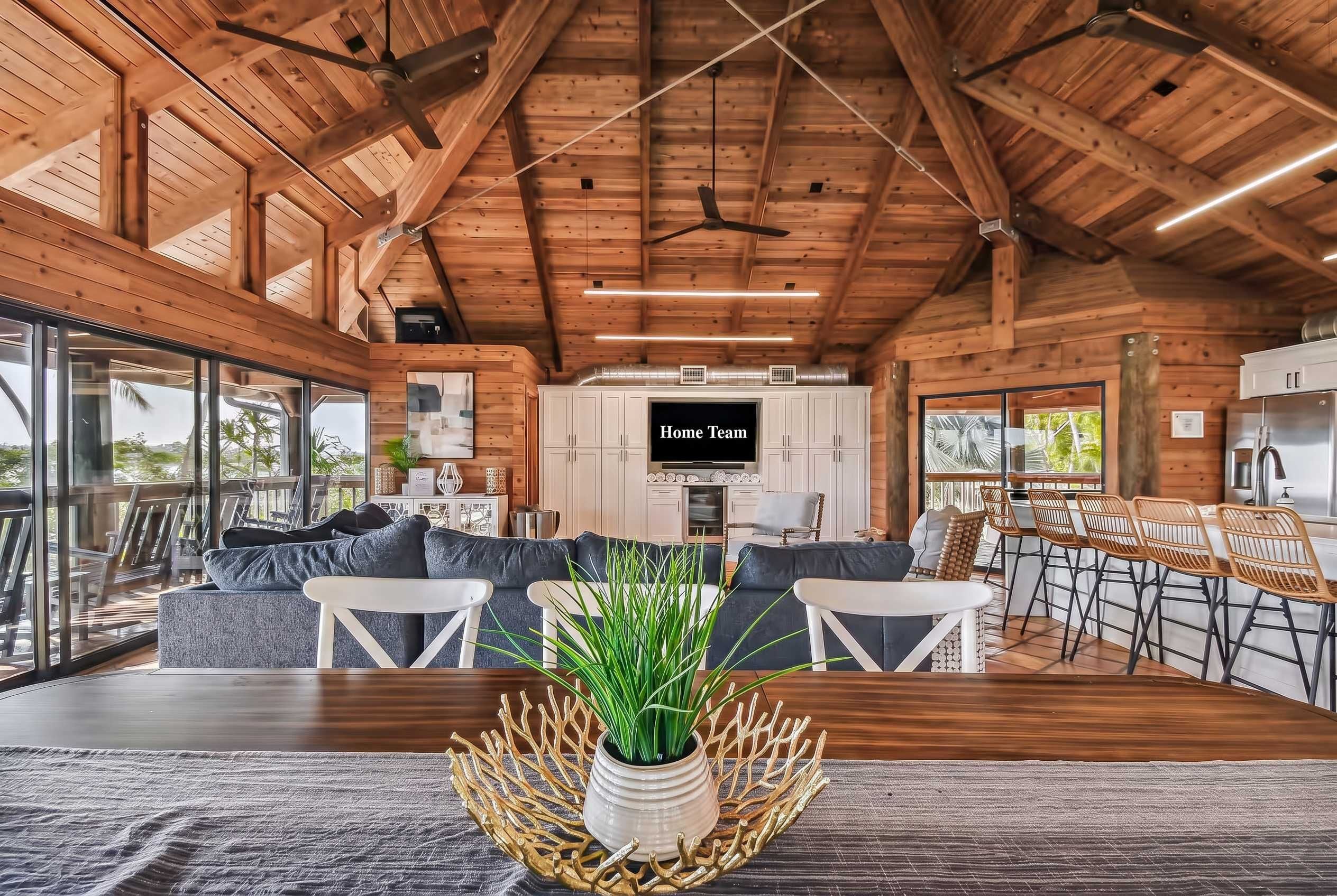 A spacious living area with a wooden ceiling, a dining area with a bar, and a kitchen with a TV labeled 'Home Team'.