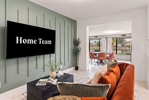 A modern living room with a flat-screen TV, a coffee table with magazines, a sofa with cushions, and a dining area visible through a glass door. - Lakin It Easy Luxe Waterfront Oasis Sleeps 16 (Mesa)