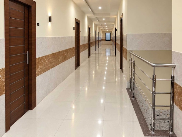 Un pasillo largo y bien iluminado con piso pulido, una puerta de madera a la izquierda y una barandilla metálica a la derecha.