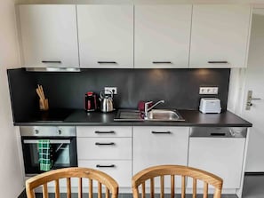 A modern kitchen with white cabinets, a black countertop, a kettle, a toaster, and a sink. There are two wooden chairs and a green towel hanging in the oven. - Dhow Flat at Paje Beach | Step from your Garden into the Pool (Paje)
