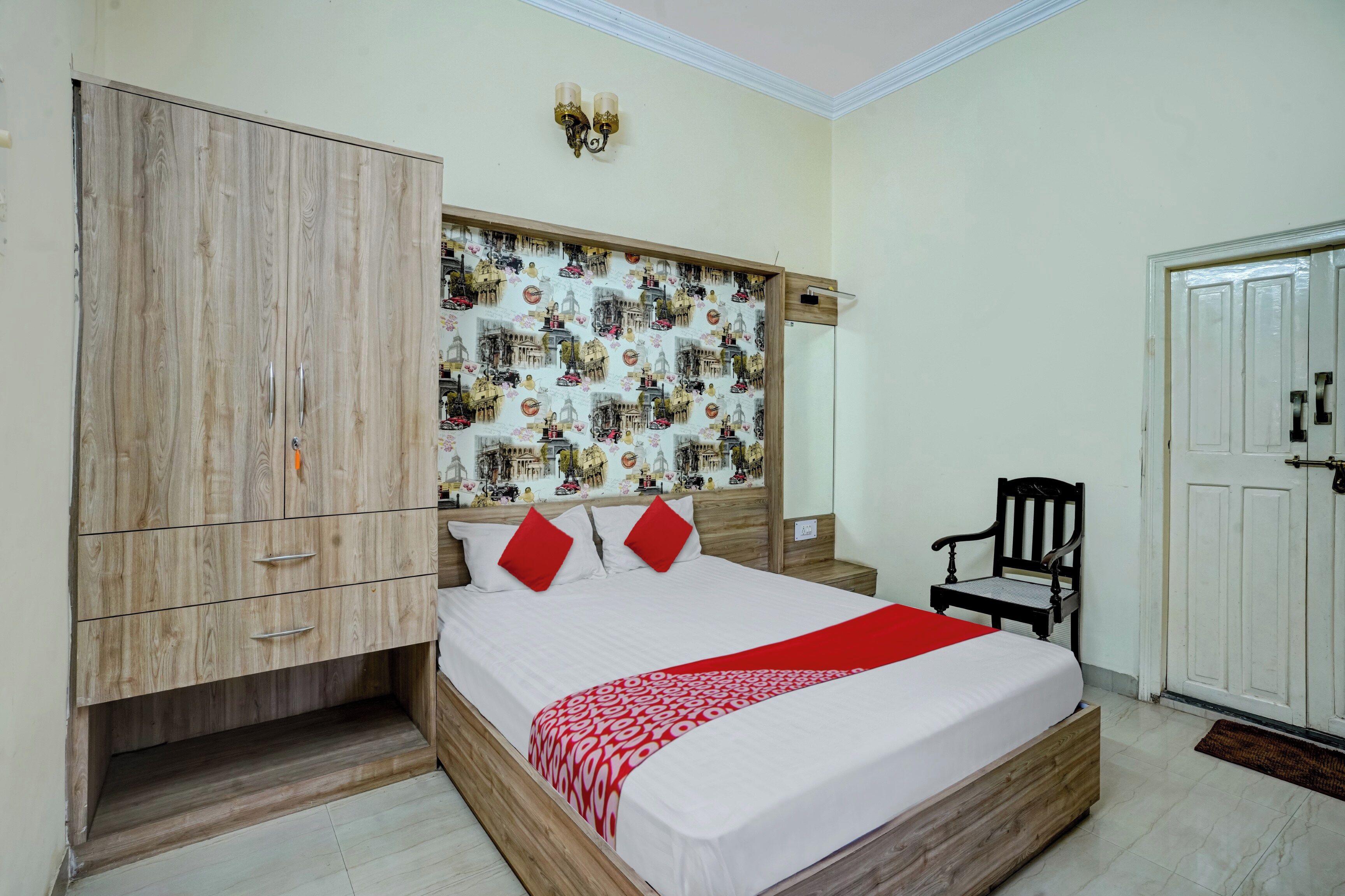 A bedroom with a wooden wardrobe, a bed with red pillows, a chair, and a wall with a cityscape mural.