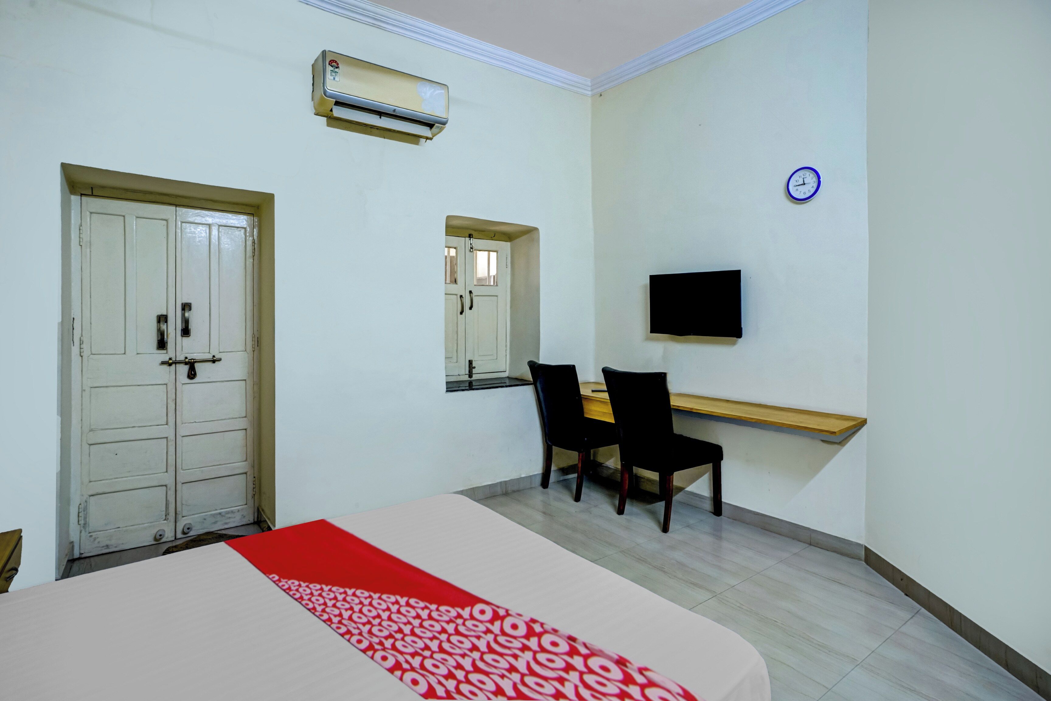 A hotel room with a bed, a desk with two chairs, a flat-screen TV, an air conditioning unit, and a clock on the wall.