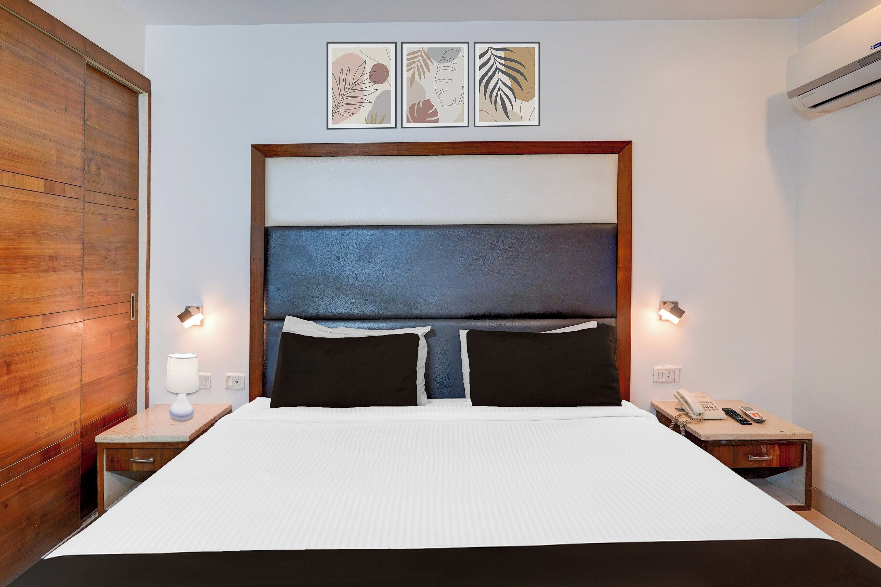 A hotel room with a large bed, two bedside tables, a wooden wardrobe, and three framed artworks on the wall.