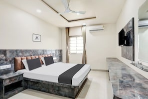 Une chambre d’hôtel comprenant un lit, une table de chevet, une commode, un ventilateur de plafond, un appareil de climatisation, une fenêtre avec des rideaux et un téléviseur fixé au mur.