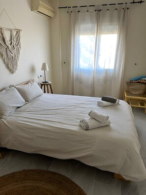 A neatly made bed with white linens and towels, a small bedside lamp, a macrame wall hanging, and a window with sheer curtains. - Apartment 'Casa Kapano' with Mountain View, Wi-Fi and Air Conditioning (La Cala de Mijas)