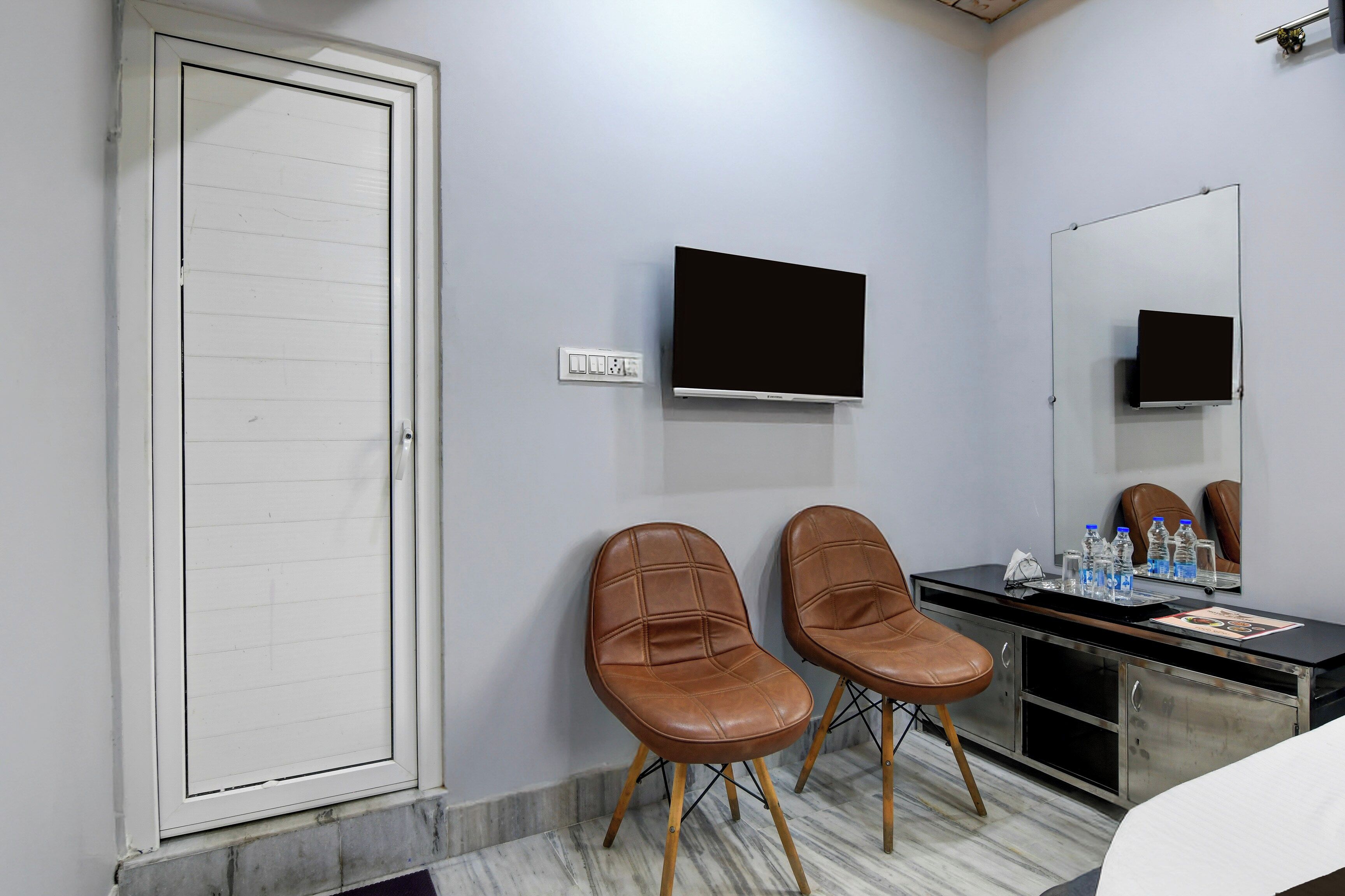 Habitació d'hotel amb una porta blanca, un televisor de pantalla plana fixat a la paret, dues butaques de pell marró, un escriptori amb mirall i una nevera minibar.