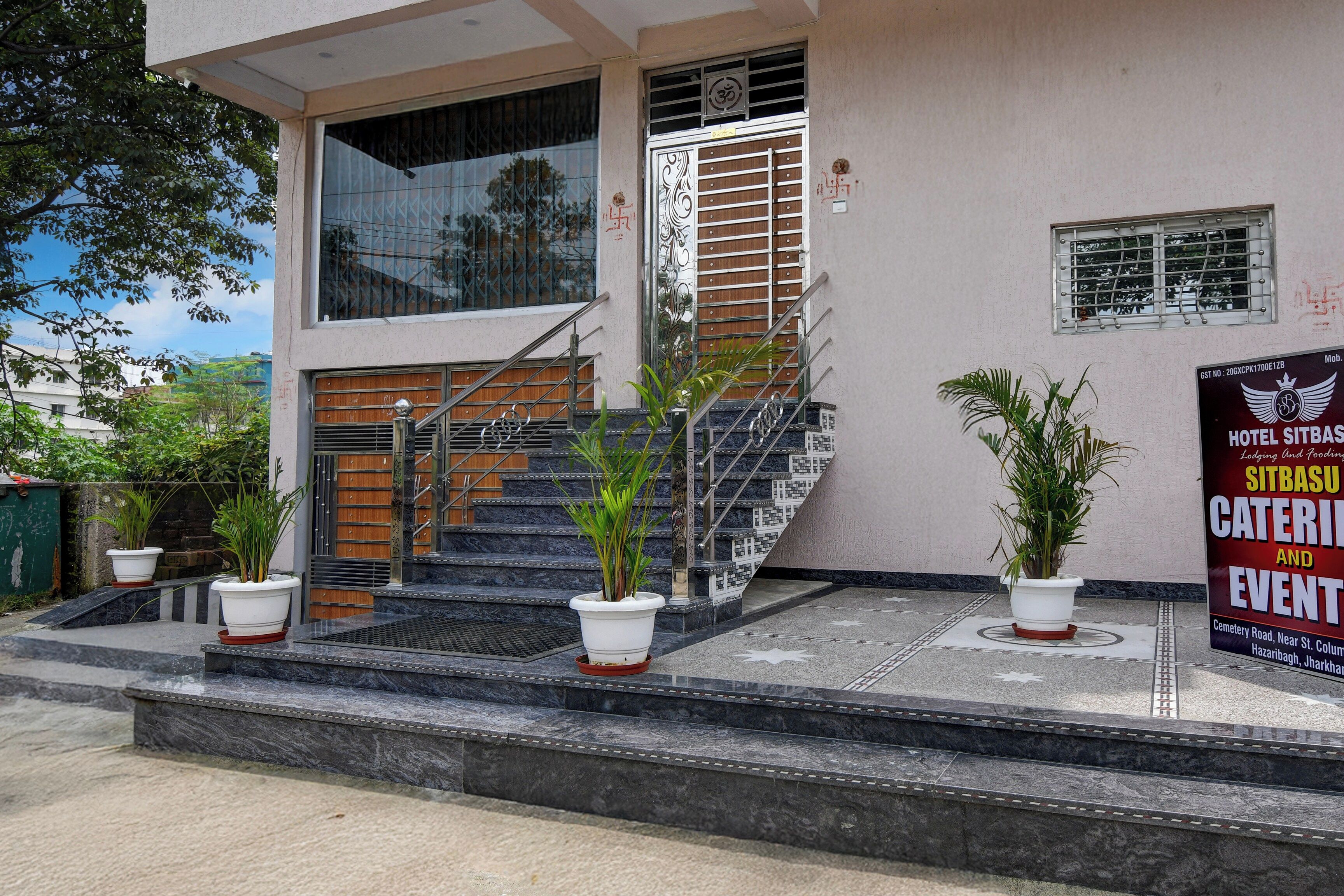 Entrada de hotel con escaleras, plantas en macetas y un cartel anunciando servicios.