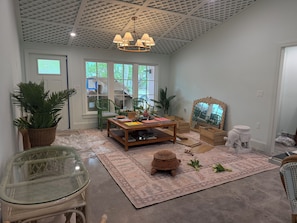 A room with a wooden coffee table, green chairs, a glass table, a woven basket, and a large mirror. - Spoonbill Cottage | 3-Min Walk to Beach | Heated Pool | 4BR Family Retreat (Bradenton Beach)