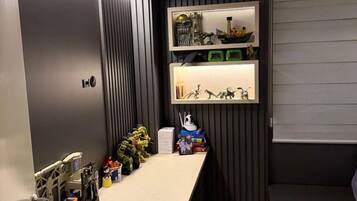 Une pièce avec un bureau, des étagères remplies de jouets et une fenêtre avec des stores.