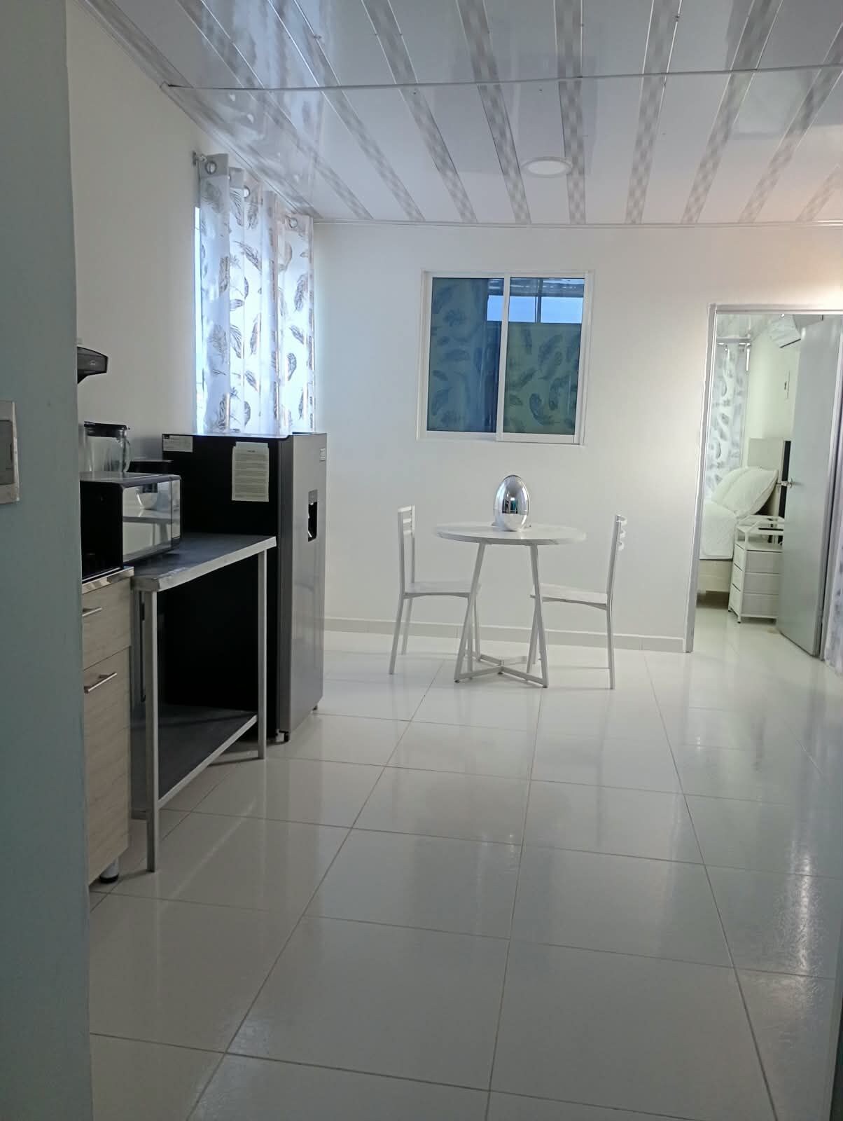 Una cocina limpia y moderna con comedor, pisos de baldosas blancas y un ventanal con cortinas.