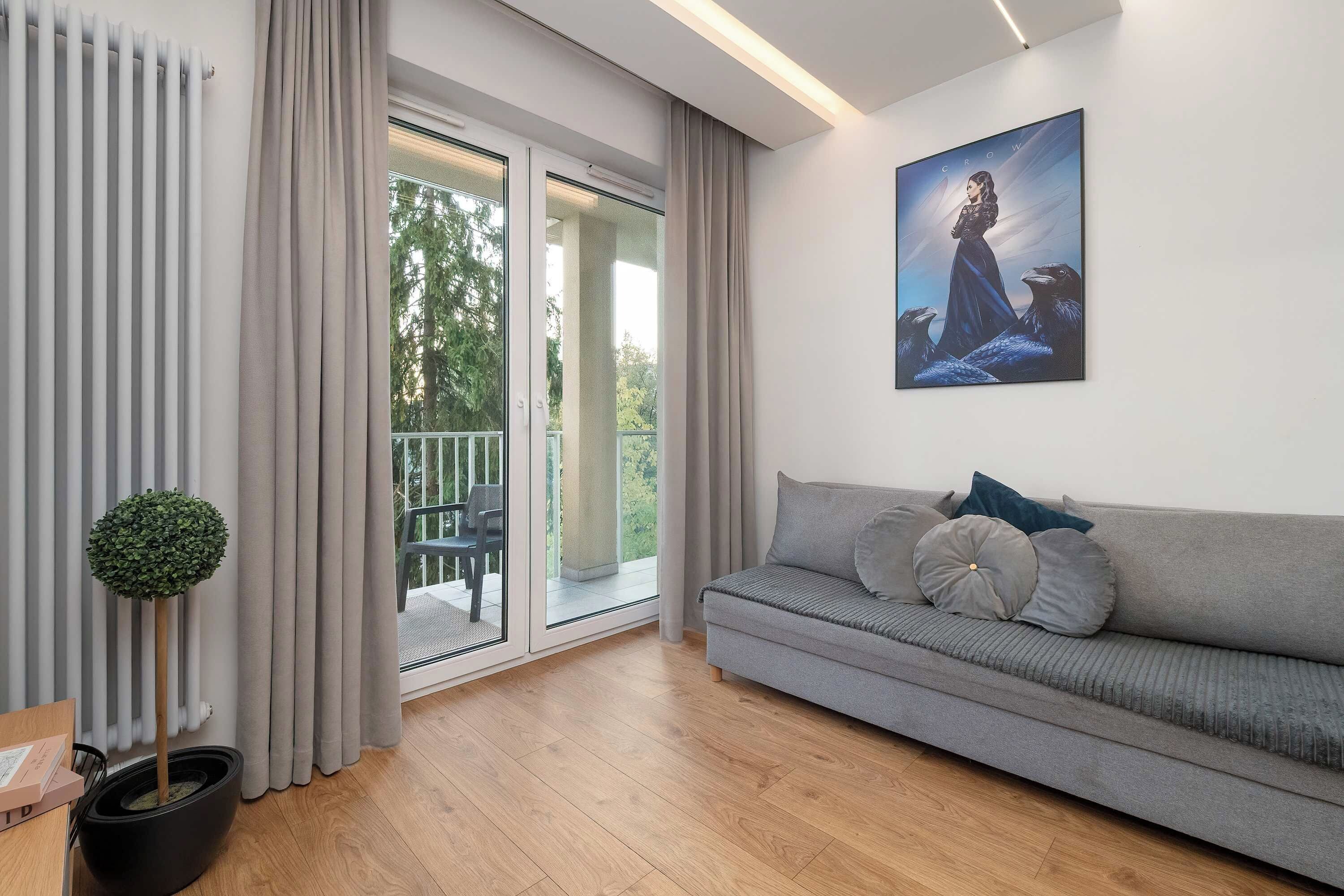 Uma sala de estar moderna com um sofá cinzento, um quadro na parede e vista para área externa através de uma porta de vidro.