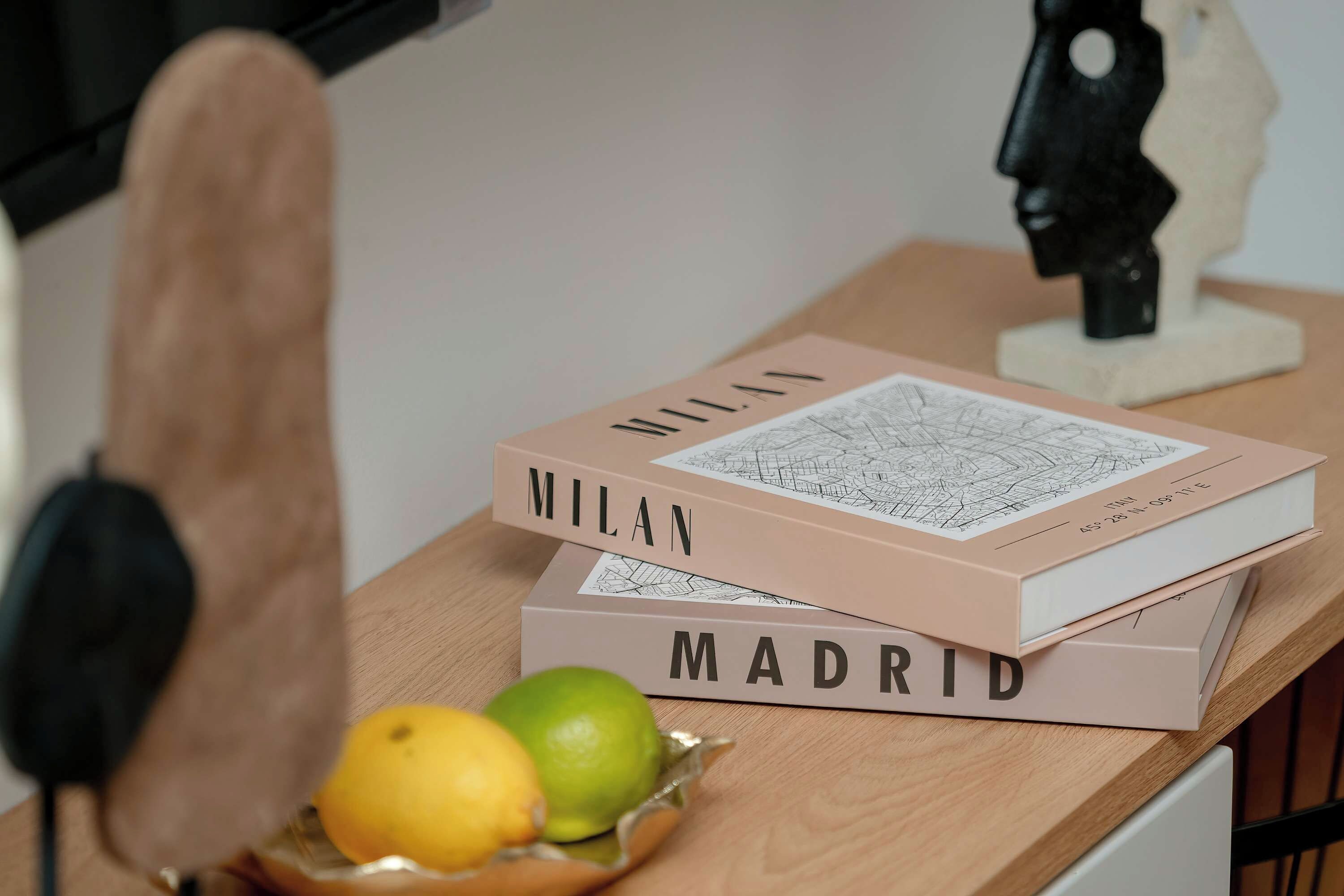 A stack of books titled 'MILAN' and 'MADRID' on a wooden surface with a pair of headphones and a fruit bowl nearby.