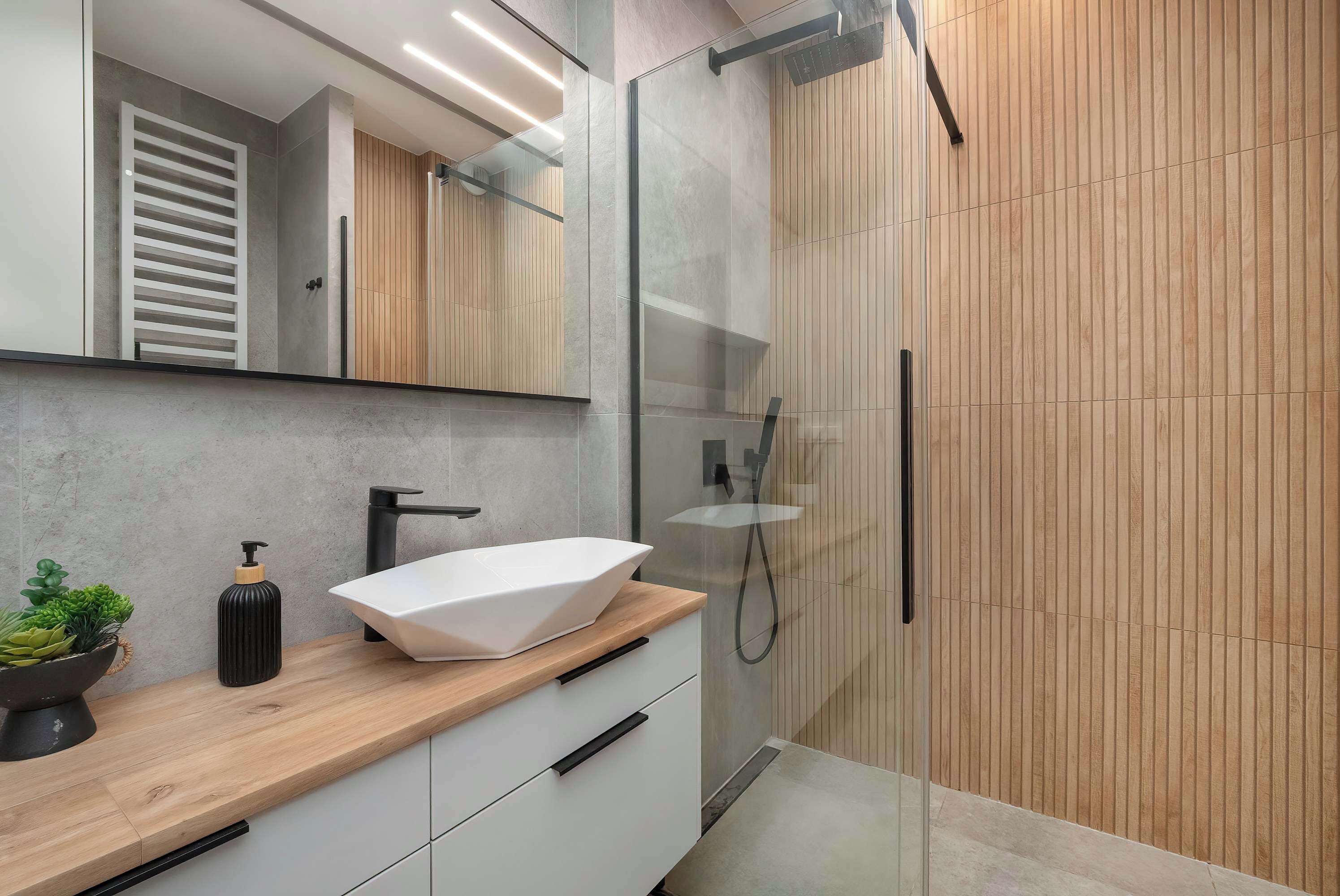 Bathroom showcasing a stylish white vessel sink atop a natural wood vanity underneath an illuminated mirror. The walk-in shower features modern glass panels and warm wood-effect tiling.