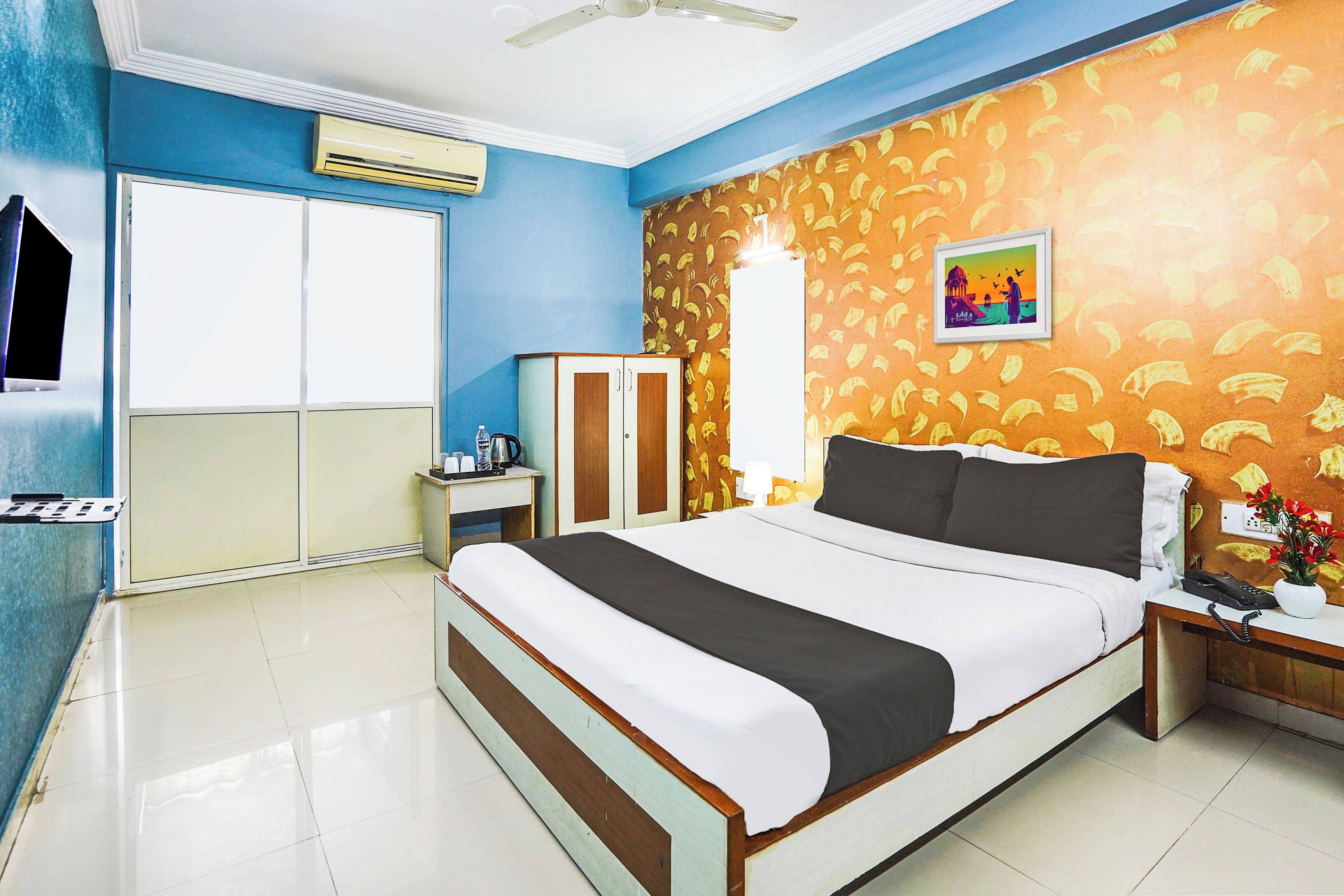 A hotel room with a bed, a wardrobe, a desk with a chair, a nightstand, a telephone, and a wall-mounted air conditioner.