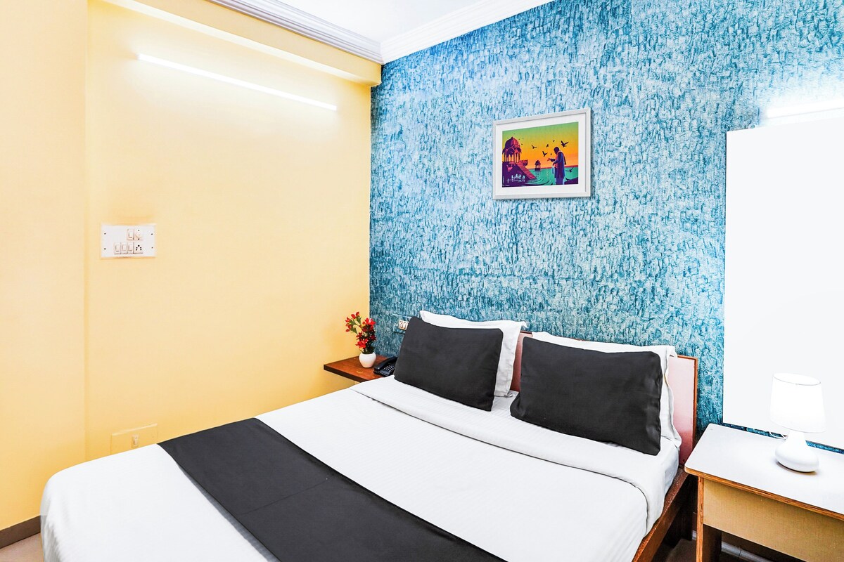 Habitación de hotel con cama, mesita de noche, un florero con flores y un cuadro en la pared.