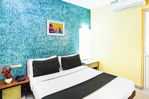 A hotel room with a bed, a nightstand, a lamp, a picture on the wall, and a wall-mounted air conditioner. - Super Hotel O Vibrant Residency (Ahmedabad)