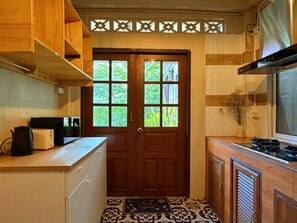 A kitchen with wooden cabinets, a black kettle, a white toaster, a stove, and a window with a view of greenery. - Tranquil 2-bedroom house with AC in charming Wat Ket Chiang Mai (Chang Wat Chiang Mai)
