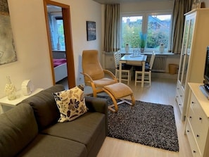Ein gemütlicher Wohnbereich mit einer Couch, einem Sessel, einem Esstisch mit Stühlen, einem Fernseher und einem Fenster mit Vorhängen.