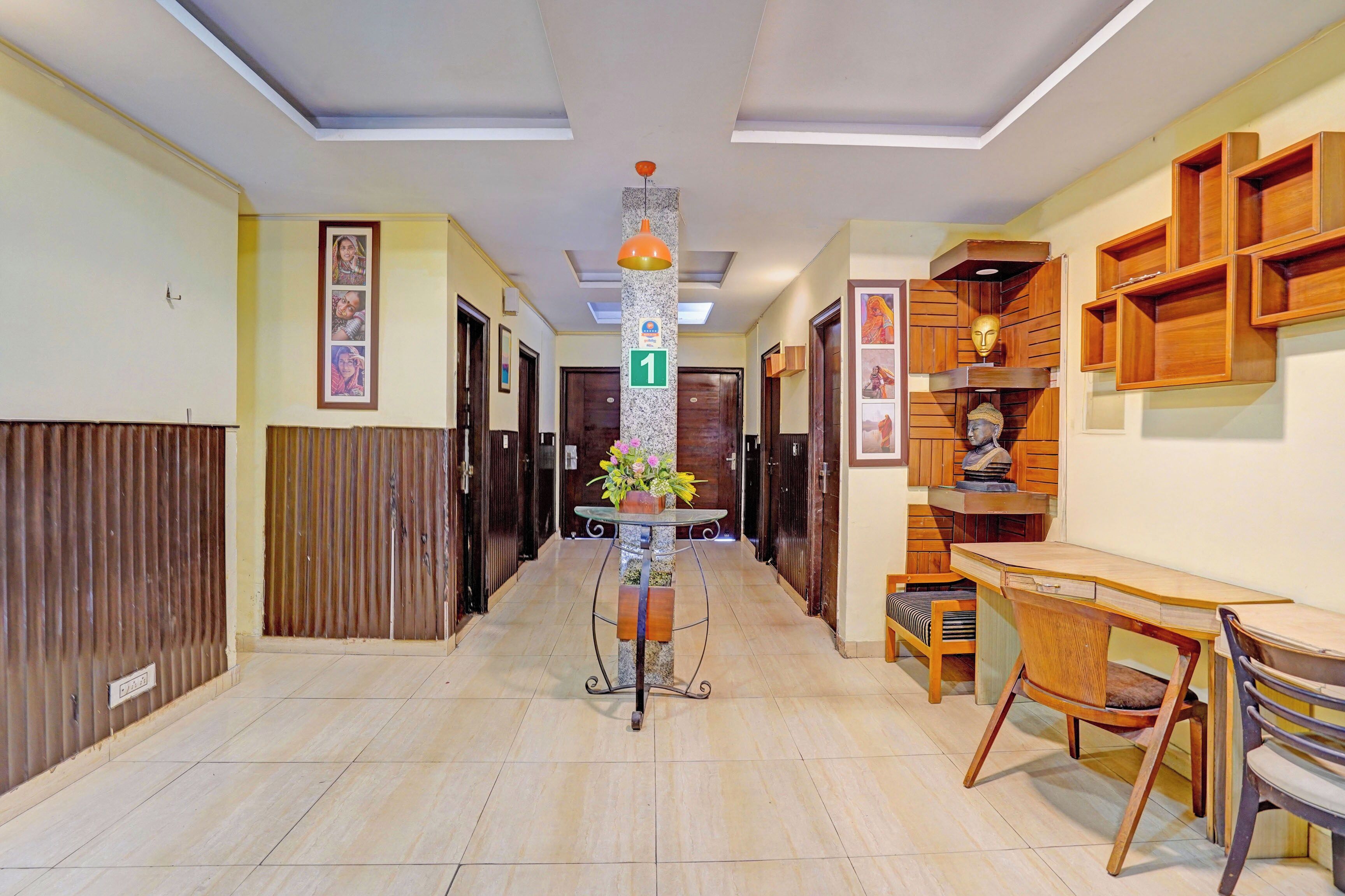 A hallway with a tiled floor, a glass table with a flower vase, a wooden desk with chairs, and a wall with wooden shelves and framed pictures.