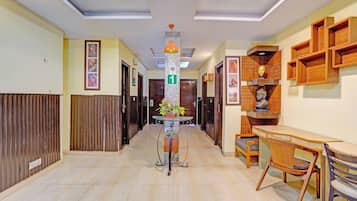 A hallway with a tiled floor, a glass table with a flower vase, a wooden desk with chairs, and a wall with wooden shelves and framed pictures.