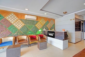 Uma área de recepção com design moderno, incluindo balcão de atendimento, espaço de espera com cadeiras de vime e uma parede decorativa com padrões geométricos.