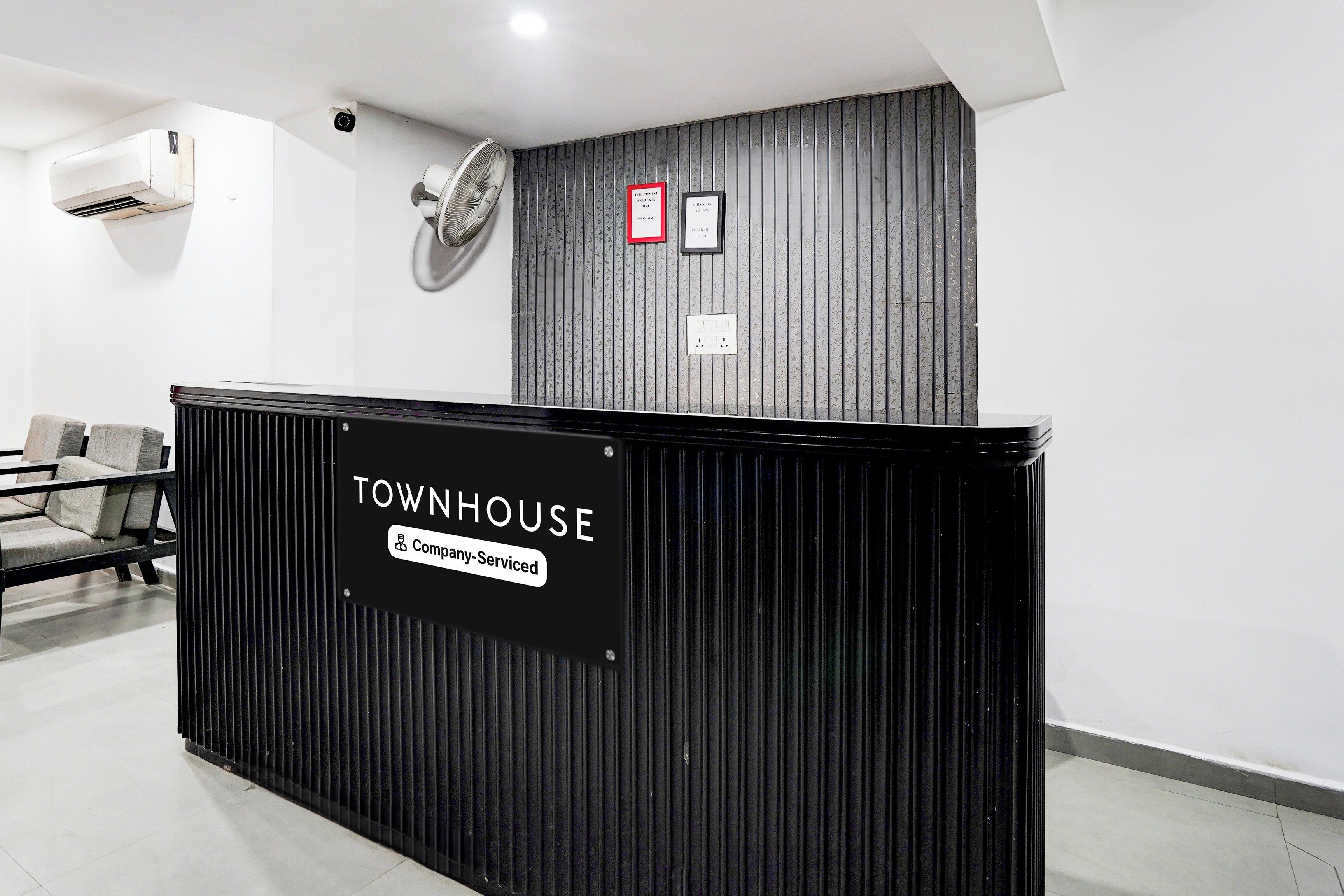 Un mostrador de recepción con la etiqueta 'TOWNHOUSE', un cartel que indica 'Servicio de Empresa', una cámara de seguridad, un detector de incendios y un ventilador de pared.