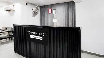 Un mostrador de recepción con la etiqueta 'TOWNHOUSE', un cartel que indica 'Servicio de Empresa', una cámara de seguridad, un detector de incendios y un ventilador de pared.
