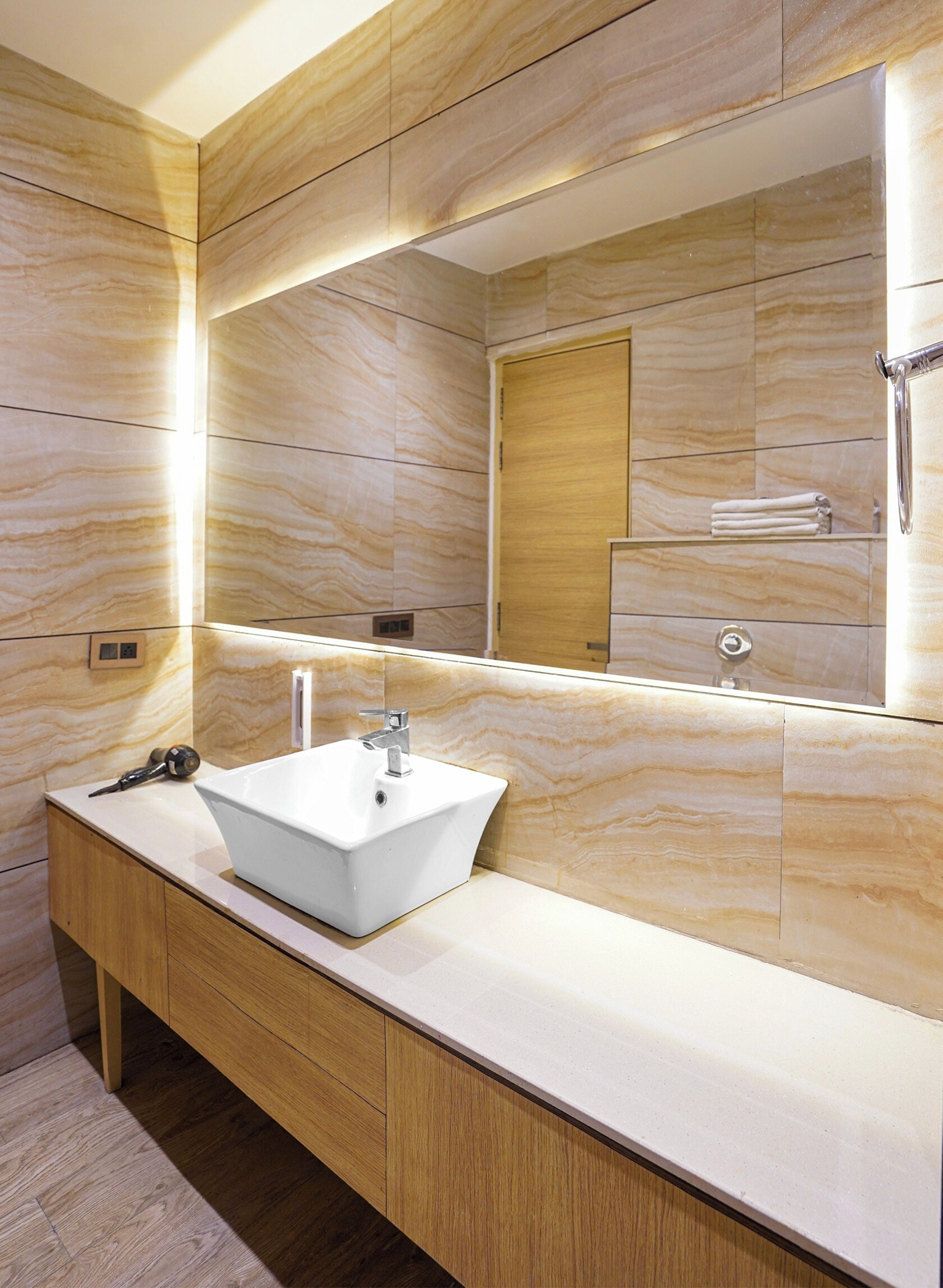Un baño con un lavamanos blanco, tocador de madera, paredes de mármol y un espejo grande.
