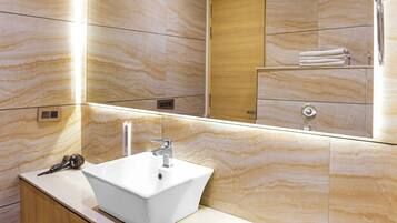Un baño con un lavamanos blanco, tocador de madera, paredes de mármol y un espejo grande.