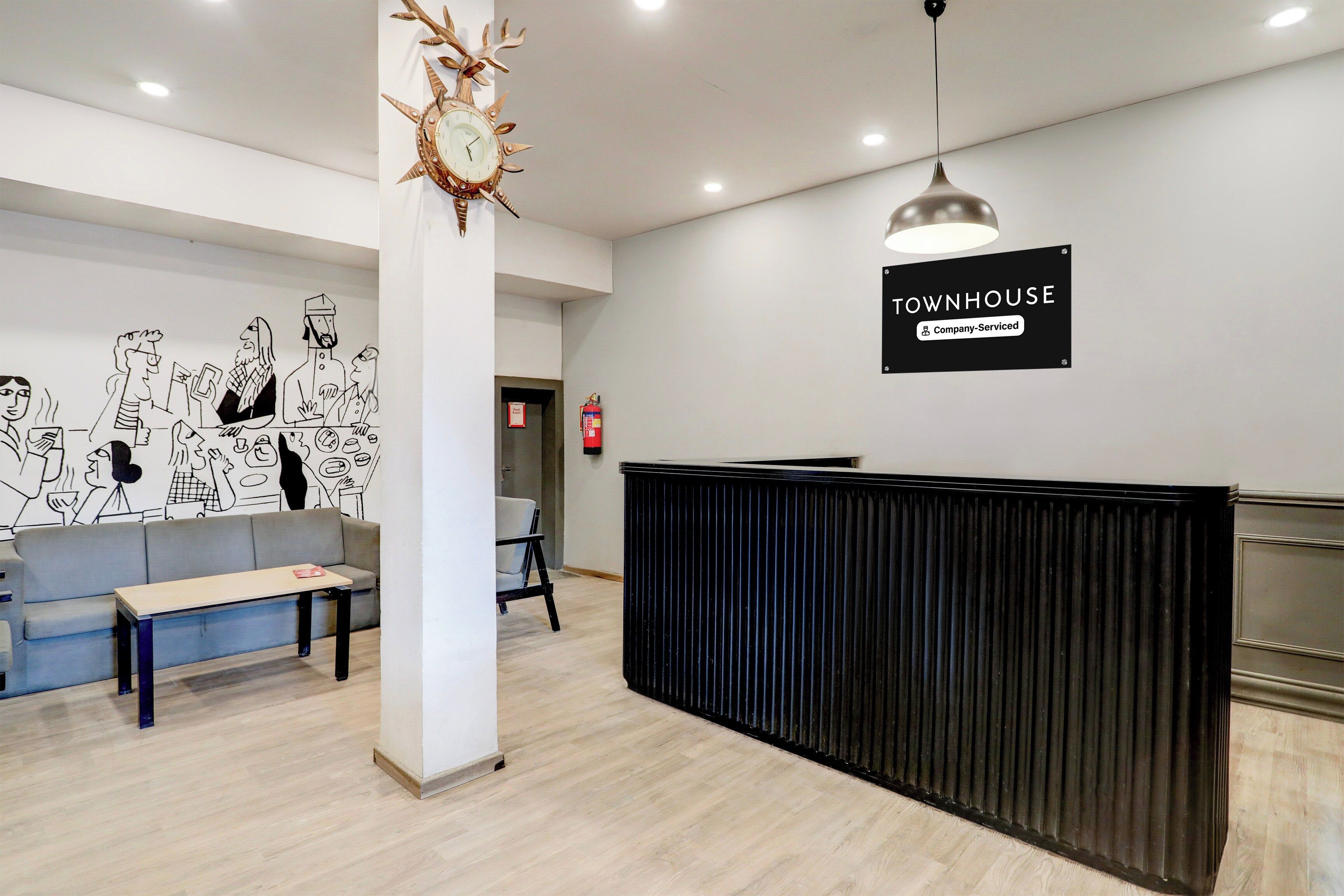 A reception area with a black reception desk, a wooden clock, a grey sofa, and a wall with a black and white illustration.