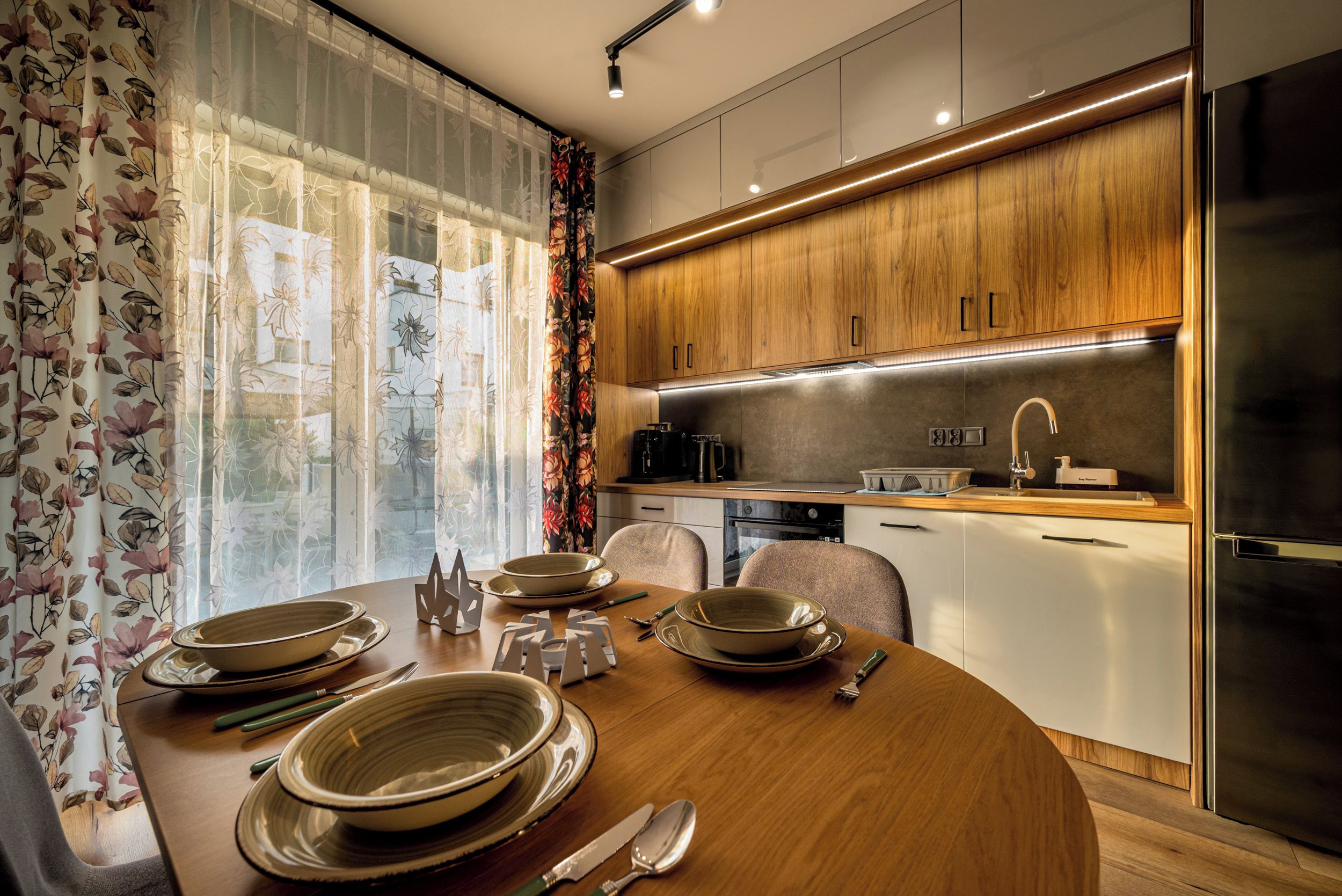 A modern kitchen with wooden cabinets, a stainless steel refrigerator, and a dining table set with plates and cutlery.