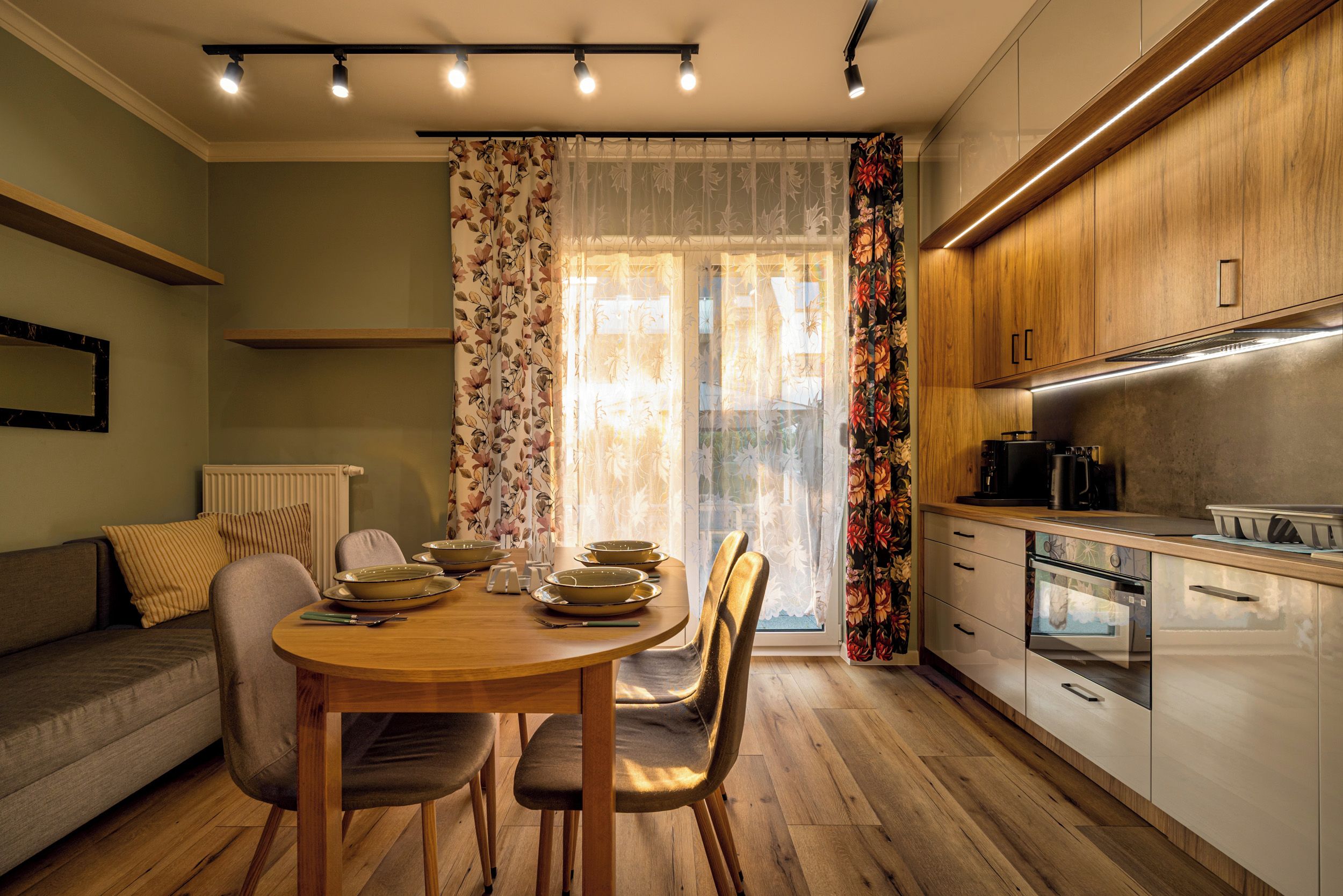 A modern kitchen with wooden cabinets, a dining area with a table and chairs, and a window with floral curtains.