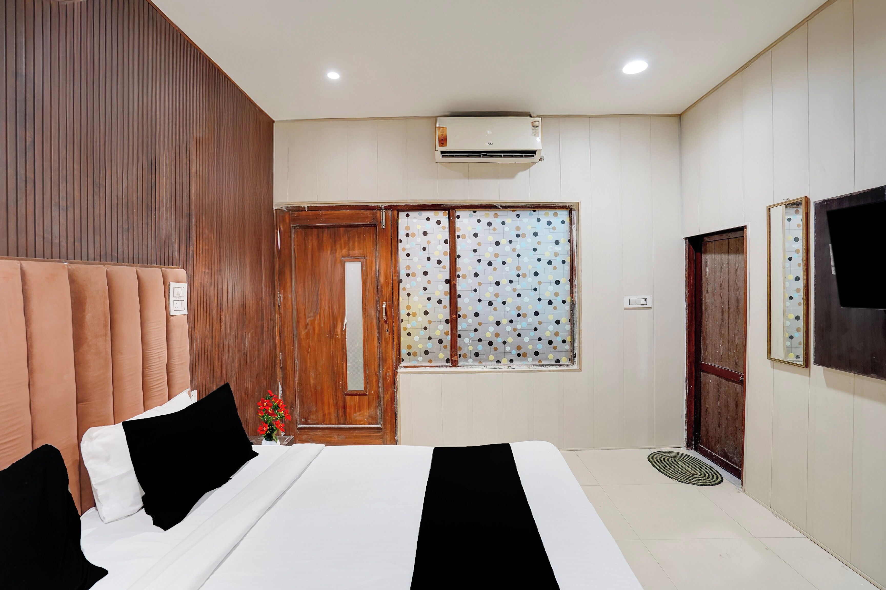 Habitación de hotel con cama, cabecera de madera, ventana con persiana de lunares, unidad de aire acondicionado y televisor montado en la pared.
