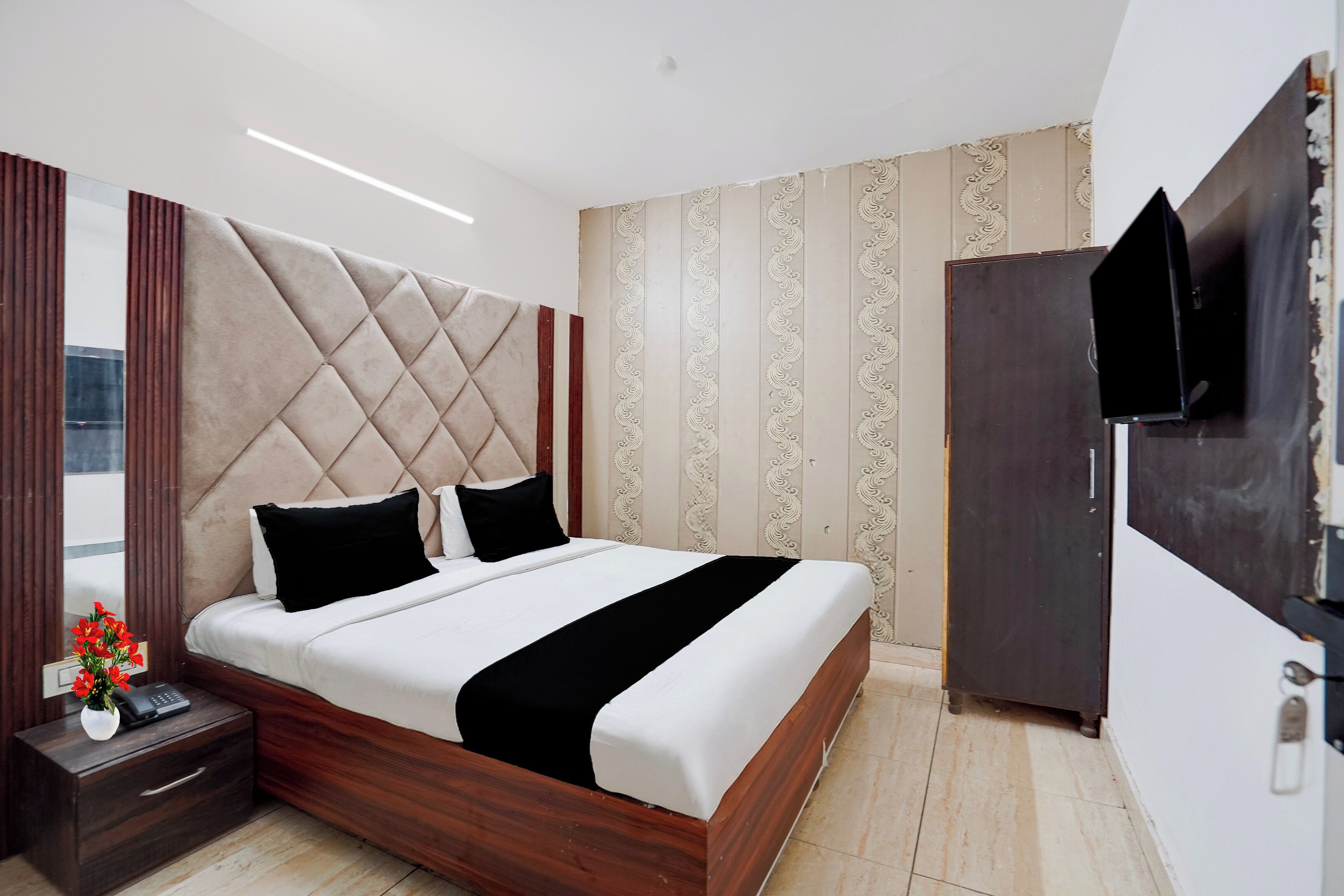 A modern bedroom with a tufted headboard, a wooden bed frame, a bedside table with a vase of flowers, and a TV mounted on the wall.