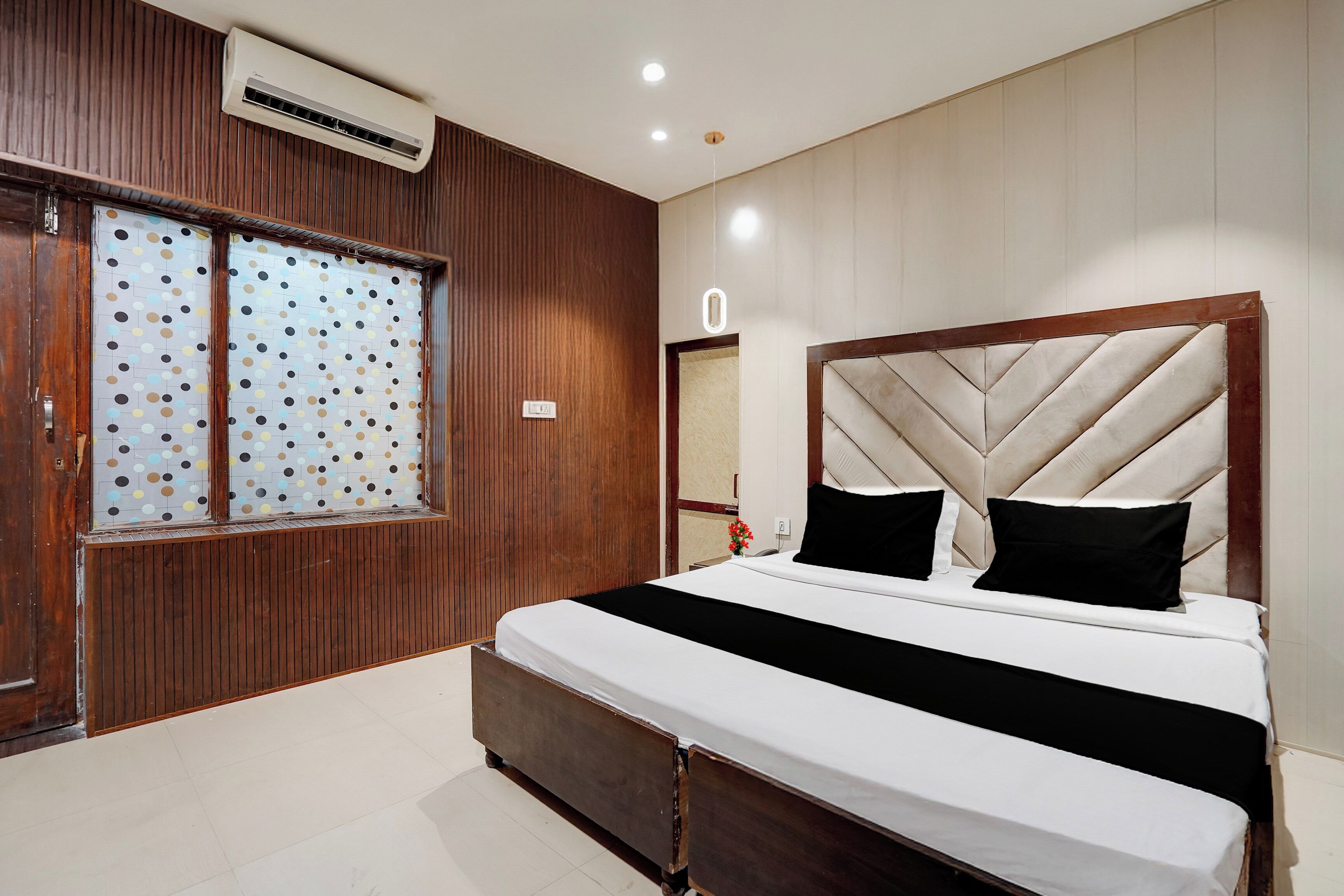 Quarto com cama, painéis de madeira, janela com padrão de poços e um aparelho de ar condicionado.