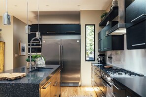A modern kitchen with stainless steel appliances, wooden cabinetry, and a large window providing natural light. - Jacaranda House | Designer Retreat with Private Hot Tub (Venice, Los Angeles)