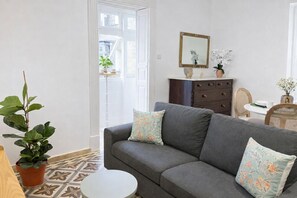 A living room with a grey sofa, two patterned cushions, a small round table, a potted plant, a dining area with a table and chairs, and a wall-mounted mirror. - Nuraghi (Il-Furjana)