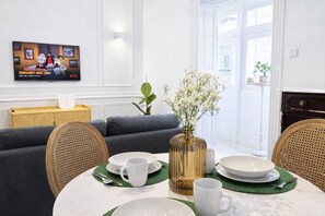 A dining area with a round table set for two, a television mounted on the wall, and a sofa in the background. - Nuraghi (Il-Furjana)