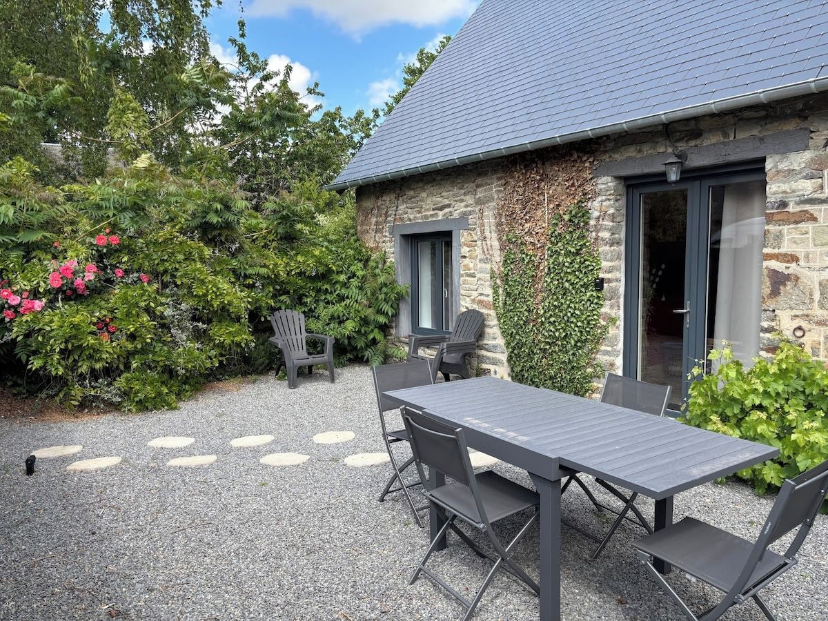Ein Steinhaus mit grauem Dach, einer Schotterterrasse mit Tisch und Stühlen sowie einem Garten mit Büschen und Blumen.