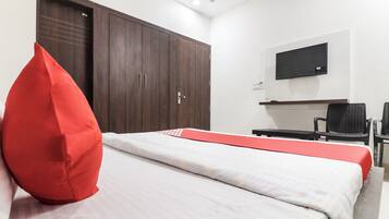 A hotel room with a bed, a red pillow, a TV mounted on the wall, and a desk with chairs.