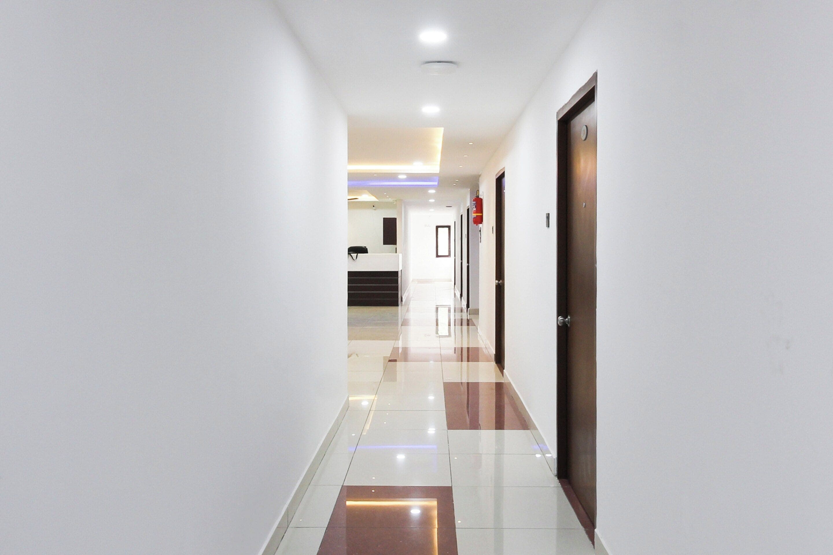 Un pasillo de hotel con diseño moderno, que presenta una combinación de piso de baldosas blancas y marrones, paredes blancas y una puerta de madera a la derecha.
