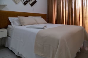 Ein ordentlich bezogenes Bett mit weißen Leinen und einer beige farbenen Tagesdecke, ein hölzerner Kopfseitenrahmen, ein weißer Nachttisch und eine an der Wand montierte Uhr.