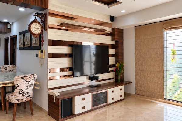 Un salon moderne doté d’un téléviseur à écran plat, d’un mur recouvert de panneaux en bois et en blanc, d’un coin repas avec une chaise à motifs papillons et d’un petit réfrigérateur.