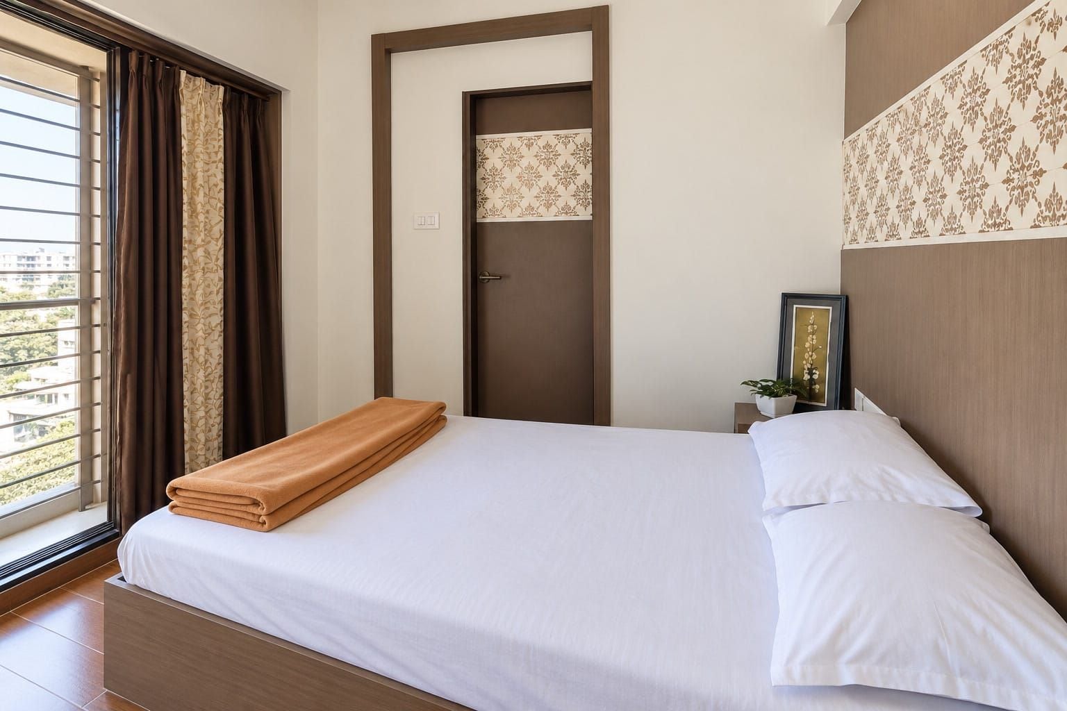Ein Hotelzimmer mit Bett, einem Fenster mit Vorhängen, einer Tür und einem kleinen Nachttisch mit Pflanze.