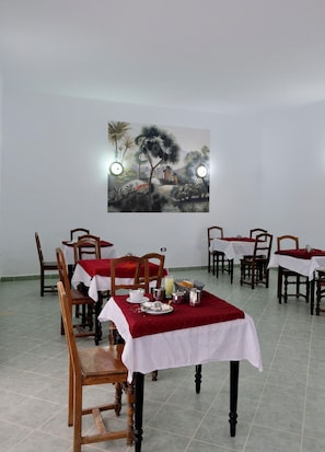 Uma área de refeições com mesas para dois, cadeiras e um quadro pendurado na parede.