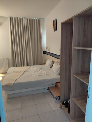 Quarto com cama, mesa de cabeceira, guarda-roupa e janela com cortinas.