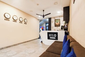 Zona de recepción de un hotel con un mostrador, un sofá y relojes que muestran la hora de Nueva Delhi, Nueva York, Londres y Tokio.
