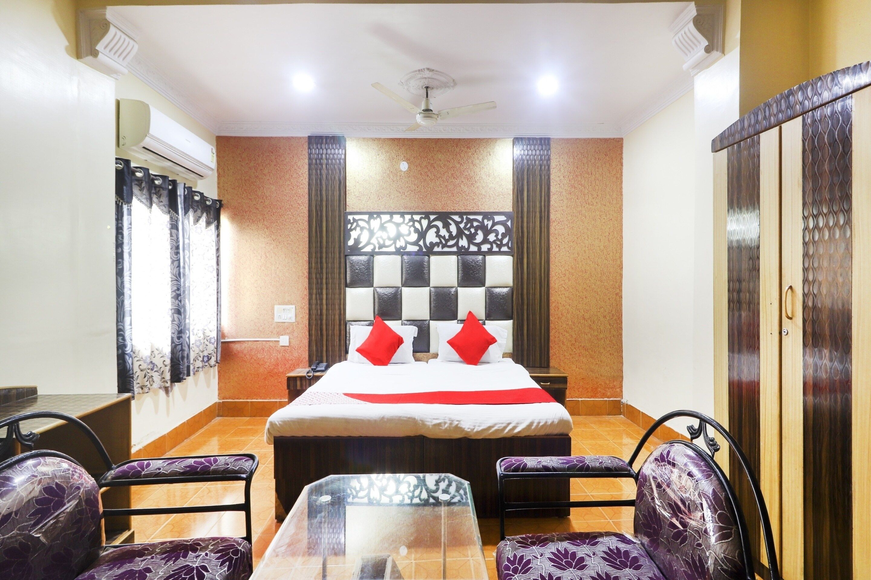 Une chambre d’hôtel comprenant un lit, deux chaises, une petite table, un ventilateur de plafond et un panneau décoratif mural.