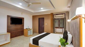 A hotel room with a bed, a TV mounted on the wall, a ceiling fan, a small bedside table, and a potted plant.